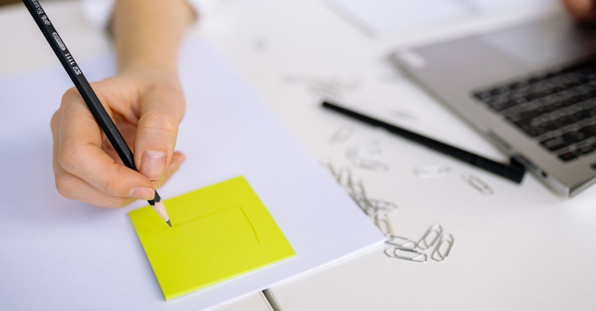 Close-up of hand sketching on a sticky note beside a laptop with paper clips on a white table.